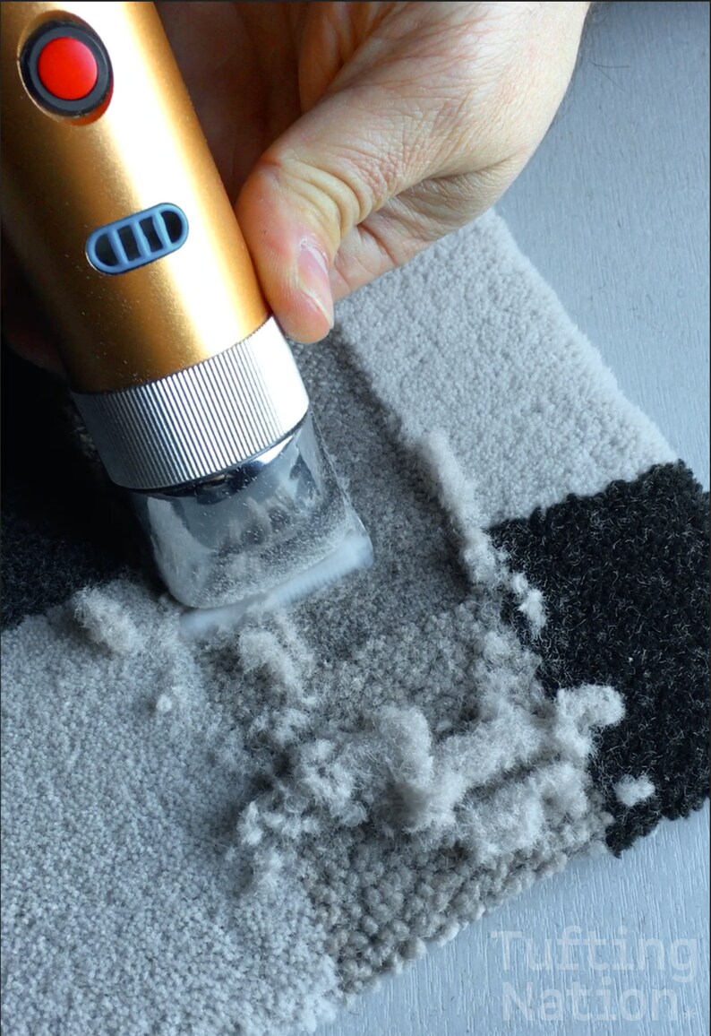 May include: A close-up of a hand using a handheld electric fabric shaver to remove pilling from a gray and black rug. The shaver has a gold body and a silver head.