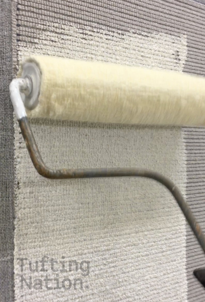 Op de afbeelding: Een close-up van een tapijt dat wordt bewerkt met een verfroller. De roller is cr&egrave;mekleurig en het tapijt is een mix van grijs en cr&egrave;me. De tekst "Tufting Nation" is zichtbaar onderaan.