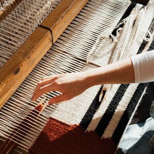 K&ouml;nnte beinhalten: Eine Hand webt einen Teppich auf einem h&ouml;lzernen Webstuhl. Der Teppich besteht aus wei&szlig;em, schwarzem und braunem Garn. Der Webstuhl ist aus Holz und hat einen wei&szlig;en Faden, der durch ihn l&auml;uft.
