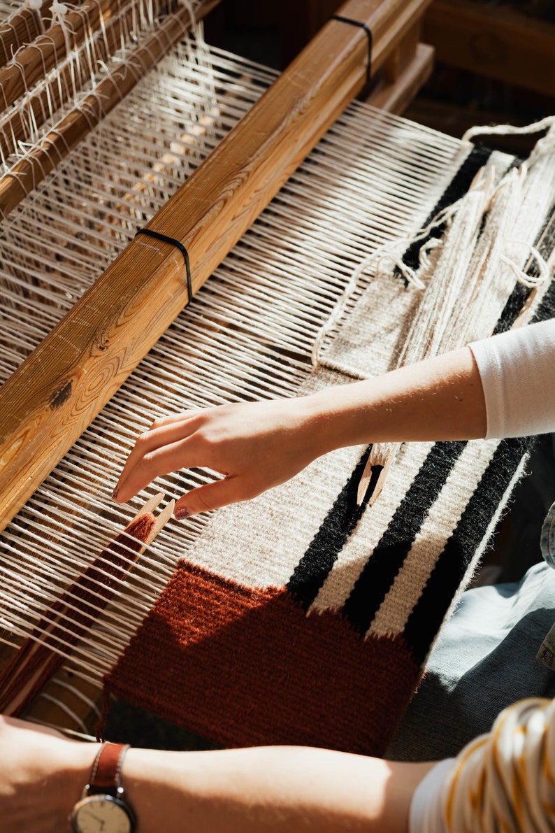 K&ouml;nnte beinhalten: Eine Hand webt einen Teppich auf einem h&ouml;lzernen Webstuhl. Der Teppich besteht aus wei&szlig;em, schwarzem und braunem Garn. Der Webstuhl ist aus Holz und hat einen wei&szlig;en Faden, der durch ihn l&auml;uft.
