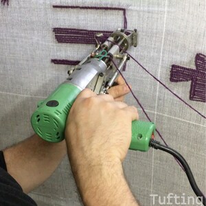 May include: A close-up of a person using a green and silver electric tufting gun to create a purple design on a gray fabric. The gun has a cord and is held in the person's right hand. The fabric is stretched taut on a frame.