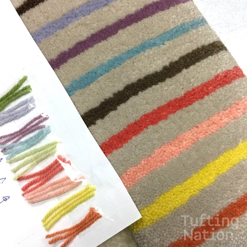 K&ouml;nnte beinhalten: Nahaufnahme eines Teppichs mit einem gestreiften Muster in verschiedenen Farben, darunter Gr&uuml;n, Lila, Blau, Orange, Gelb und Rosa. Der Teppich wird neben einer Musterkarte mit Garn in passenden Farben gezeigt.