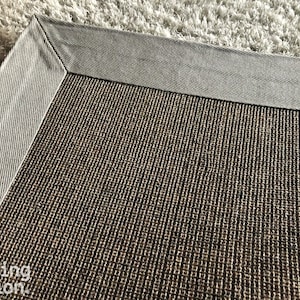 Op de afbeelding: Close-up van een rechthoekig tapijt met een getextureerd, bruin midden en een grijze rand. De hoek van het tapijt is zichtbaar, met de gedetailleerde weefsel en contrasterende kleuren. Het tapijt ligt op een lichtgekleurd, getextureerd oppervlak.