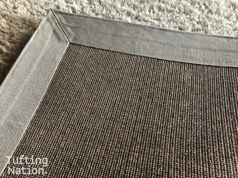 Op de afbeelding: Close-up van een rechthoekig tapijt met een getextureerd, bruin midden en een grijze rand. De hoek van het tapijt is zichtbaar, met de gedetailleerde weefsel en contrasterende kleuren. Het tapijt ligt op een lichtgekleurd, getextureerd oppervlak.