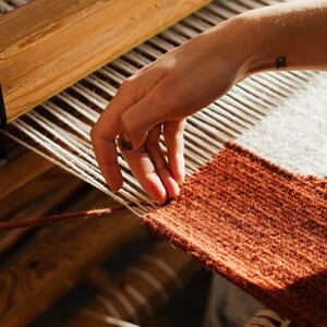 May include: A person's hand weaving a rug on a loom. The rug is made of brown, white, and black yarn. The loom is made of wood.