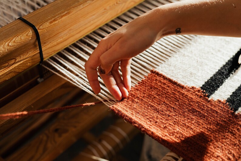 May include: A person's hand weaving a rug on a loom. The rug is made of brown, white, and black yarn. The loom is made of wood.