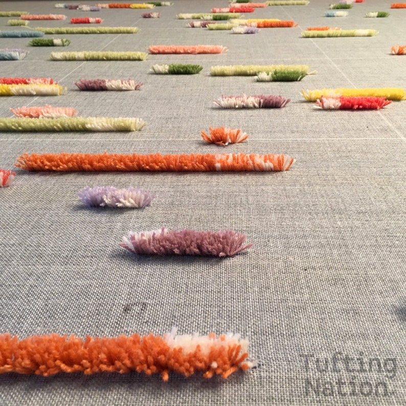 May include: A close-up of a gray fabric with colorful yarn tufts arranged in a pattern. The tufts are different lengths and colors, creating a textured and visually interesting surface.