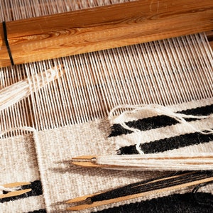 May include: A close-up of a loom with a black and white striped fabric being woven. The fabric is made with white and black yarn. The loom is made of wood and has a wooden bar at the top.