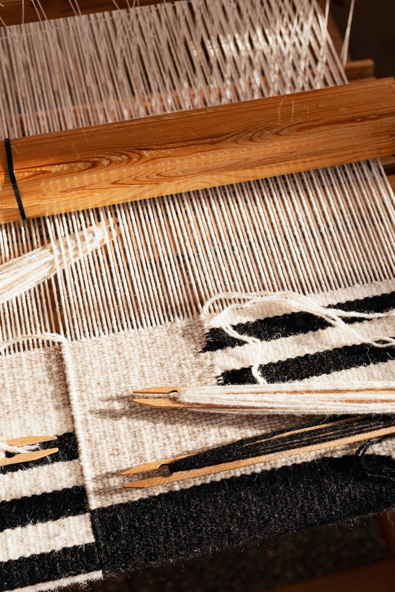 May include: A close-up of a loom with a black and white striped fabric being woven. The fabric is made with white and black yarn. The loom is made of wood and has a wooden bar at the top.