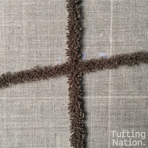 K&ouml;nnte beinhalten: Nahaufnahme eines braunen getufteten Teppichs, der auf einer beigefarbenen Leinwand hergestellt wird. Der Teppich wird in einem Kreuzmuster getuftet.