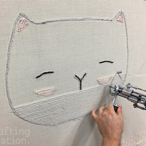 May include: A close-up of a white cat face being embroidered on a white canvas using a specialized machine. The cat has black eyes and a pink nose.