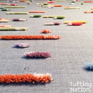 K&ouml;nnte beinhalten: Nahaufnahme eines in Arbeit befindlichen Tufting-Teppichs, der bunte Garn-Schlaufen in verschiedenen Schattierungen von Rosa, Orange, Lila, Gr&uuml;n und Gelb zeigt. Der Teppich wird auf einer grauen Stoffbasis getuftet.