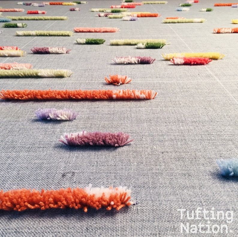 K&ouml;nnte beinhalten: Nahaufnahme eines in Arbeit befindlichen Tufting-Teppichs, der bunte Garn-Schlaufen in verschiedenen Schattierungen von Rosa, Orange, Lila, Gr&uuml;n und Gelb zeigt. Der Teppich wird auf einer grauen Stoffbasis getuftet.