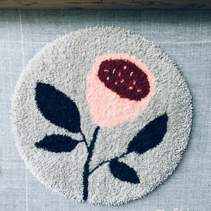 May include: A round, gray rug with a pink and red flower design. The rug is made of 100% wool and is tufted. The text "RUG TUFTING - 100% WOOL" is visible at the bottom of the image. The text "Tufting Nation" is visible at the bottom right of the image.