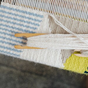 May include: A close-up of a loom with a partially woven tapestry. The tapestry features a blue and white striped pattern, a white section, and a yellow and green section. White yarn is being used to weave the tapestry.