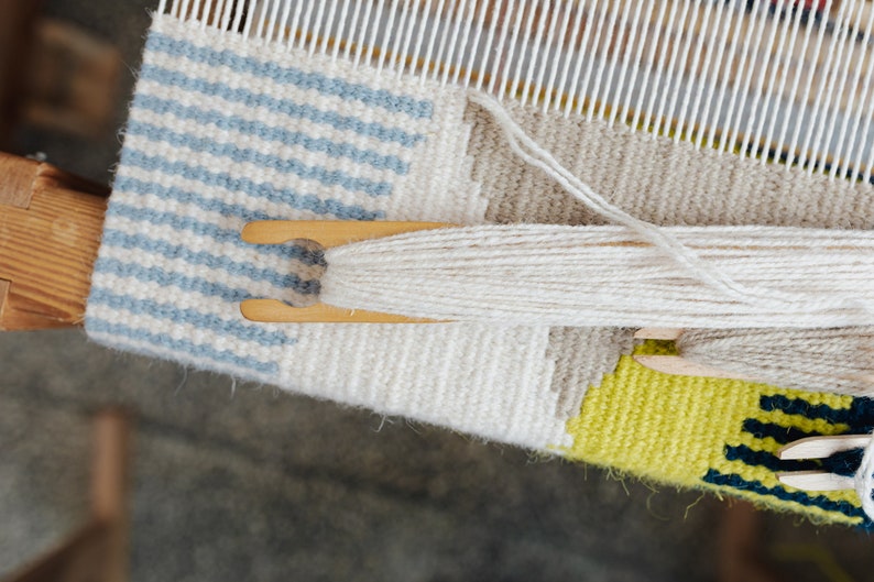 May include: A close-up of a loom with a partially woven tapestry. The tapestry features a blue and white striped pattern, a white section, and a yellow and green section. White yarn is being used to weave the tapestry.
