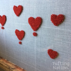 K&ouml;nnte beinhalten: Nahaufnahme einer grauen Stoffoberfl&auml;che mit sechs roten herzf&ouml;rmigen Tuften. Die Tuften bestehen aus einem flauschigen Material und sind in einem zuf&auml;lligen Muster angeordnet. Der Text "Tufting Nation" ist in der rechten unteren Ecke des Bildes sichtbar.