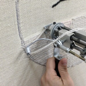 May include: A close-up of a hand holding a tufting gun, a tool used to create custom rugs and wall art. The gun is being used to create a design on a white canvas.
