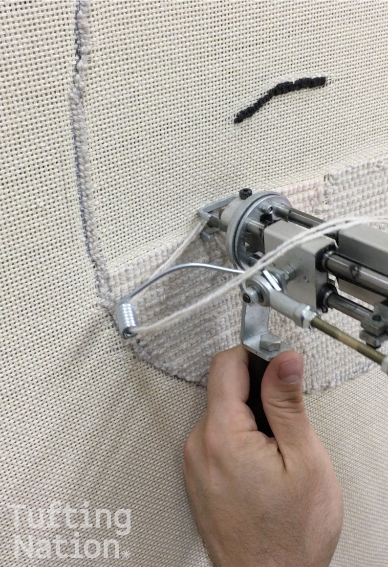 May include: A close-up of a hand holding a tufting gun, a tool used to create custom rugs and wall art. The gun is being used to create a design on a white canvas.
