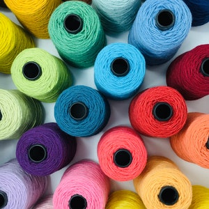 May include: A variety of colorful yarn spools in different shades of blue, green, red, yellow, orange, pink, and purple. The spools are arranged in a circular pattern on a white surface.