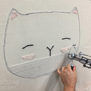 May include: A close-up of a white cat face being embroidered on a white canvas using a specialized machine. The cat has black eyes and a pink nose.