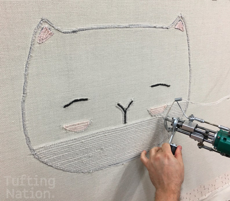 May include: A close-up of a white cat face being embroidered on a white canvas using a specialized machine. The cat has black eyes and a pink nose.