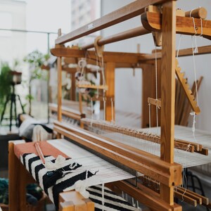 May include: A wooden loom with a black and white striped rug partially woven on it. The loom is made of natural wood and has a simple design.