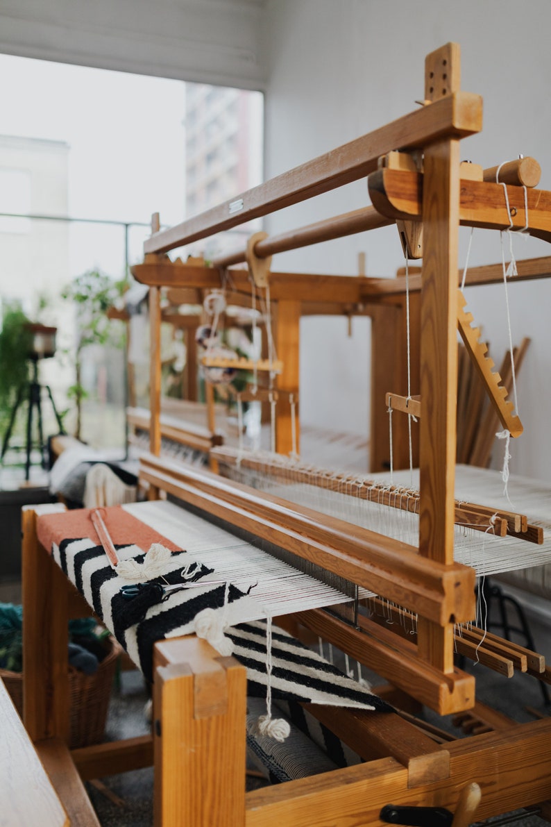 May include: A wooden loom with a black and white striped rug partially woven on it. The loom is made of natural wood and has a simple design.