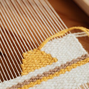 May include: Close-up of a woven tapestry on a wooden loom. The tapestry features a geometric pattern with yellow, brown, and white yarn.