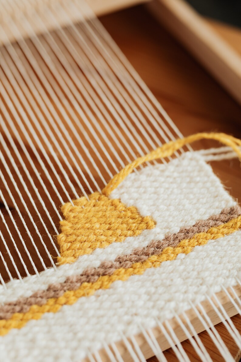 May include: Close-up of a woven tapestry on a wooden loom. The tapestry features a geometric pattern with yellow, brown, and white yarn.