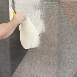 Op de afbeelding: Een persoon brengt witte lijm aan op een tapijt met een geometrisch patroon met behulp van een gele en witte spatel. Het tapijt heeft een grijs en bruin kleurenschema.