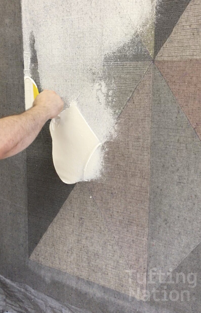 Op de afbeelding: Een persoon brengt witte lijm aan op een tapijt met een geometrisch patroon met behulp van een gele en witte spatel. Het tapijt heeft een grijs en bruin kleurenschema.