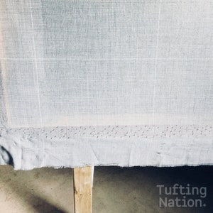 K&ouml;nnte beinhalten: Nahaufnahme eines hellgrauen Stoffes, der &uuml;ber einen Holzrahmen gespannt ist. Der Stoff hat ein Gittermuster und ist mit kleinen N&auml;geln am unteren Rand befestigt. Der Text "Tufting Nation*" ist in der unteren rechten Ecke des Bildes sichtbar.