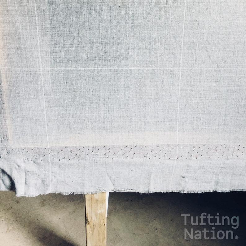 K&ouml;nnte beinhalten: Nahaufnahme eines hellgrauen Stoffes, der &uuml;ber einen Holzrahmen gespannt ist. Der Stoff hat ein Gittermuster und ist mit kleinen N&auml;geln am unteren Rand befestigt. Der Text "Tufting Nation*" ist in der unteren rechten Ecke des Bildes sichtbar.