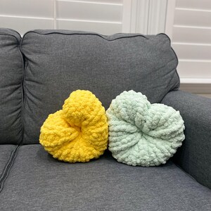 May include: Two heart-shaped decorative pillows. One is a bright yellow, the other is a soft mint green. Both pillows are made of a chunky, textured yarn and sit on a gray couch.