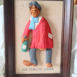 May include: A framed wood carving of a man wearing a red coat, blue scarf, and a blue beret. He is holding a bottle in his right hand. The carving is titled "Our Fearless Leader" with the artist's name "Duff Tweed" and "Pasadena" below the title.
