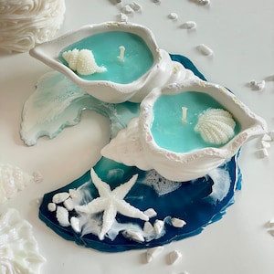 Op de afbeelding: Twee tealblauwe kaarsen in witte schelp kaarsenhouders met witte schelpen op een blauwe en witte harsbasis met een witte zeester en witte stenen.