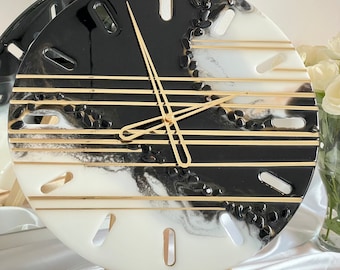 May include: A black and white resin clock with gold accents. The clock face has a marble-like design with black and white swirls and gold lines. The clock hands are gold and the clock is on a small wooden easel.
