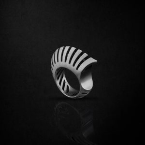 May include: A silver ring with a geometric design that resembles a sunburst. The ring is made of metal and has a smooth, polished finish.
