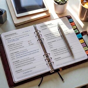 May include: A brown leather ring-bound planner open to a page titled Decision Fatigue Reducer with printed text and handwritten notes. A silver metal pen rests diagonally across the right page. The planner features colorful tabbed dividers.