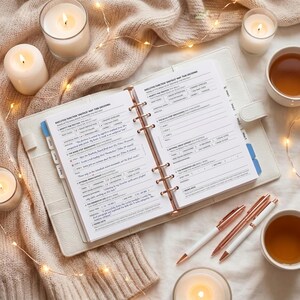 May include: A white leather planner with rose gold detailing, open to a page of printed text. The image features lit candles, two pens, and two cups of tea, arranged on a beige knitted blanket with fairy lights.