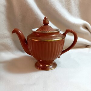 May include: A brown ceramic teapot with a gold trim and a white lid. The teapot has a ribbed design and a round body.