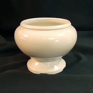 May include: A cream-colored, ceramic pedestal bowl with a rounded body and a wide, open top. The bowl sits on a short, flared base. The glossy finish reflects light, highlighting its smooth surface. The background is a dark green fabric.