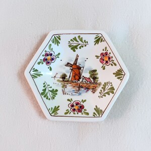 May include: A hexagonal ceramic dish with a white background and a hand-painted scene of a windmill, houses, and a canal. The dish is decorated with green leaves and pink and purple flowers.
