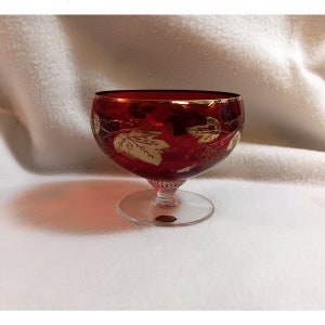 May include: A red glass goblet with a gold rim and a gold leaf design. The goblet has a clear glass stem and base.