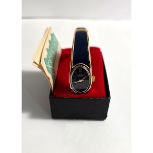 May include: A gold-tone bangle watch with a dark blue enamel inlay on the band and a matching dark blue oval dial. The watch features gold-tone hands and hour markers, a gold-tone crown, and is presented in a black box with a red velvet interior.