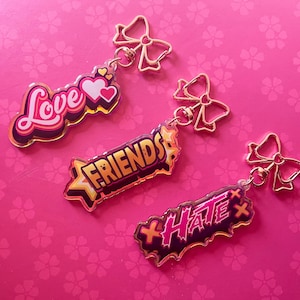 May include: Three acrylic keychains with gold bow accents. One keychain says "Love" in pink and white, another says "Friends" in yellow and orange, and the last says "Hate" in pink and black.