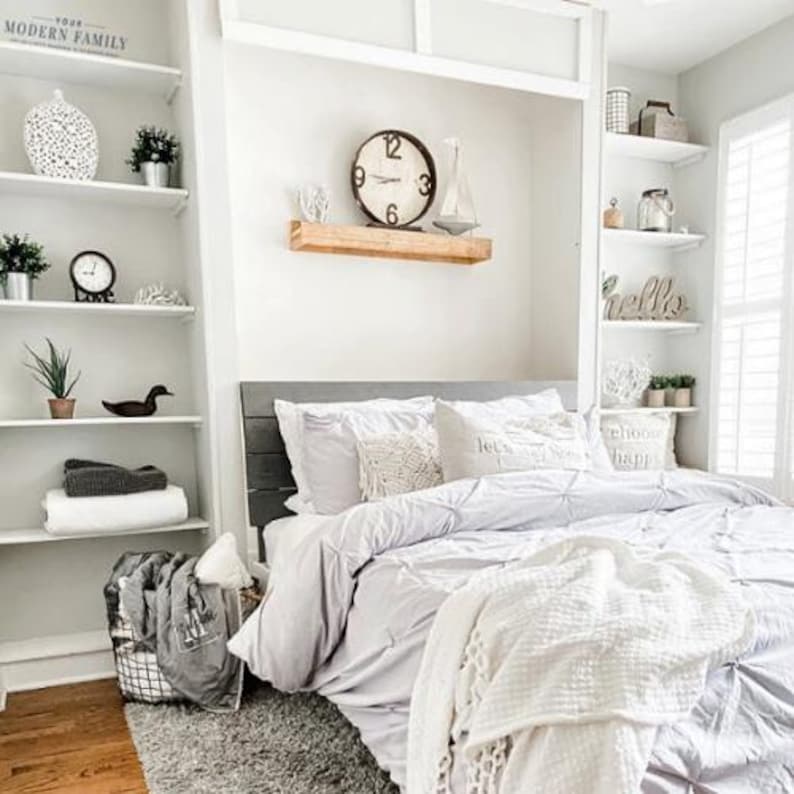 May include: A white bed with a gray headboard and a white comforter. The bed is in a bedroom with white shelves and a clock on the wall. The shelves are decorated with various items, including a white bird, a white vase, and a white clock.
