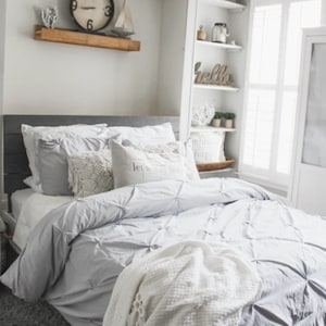 May include: A gray and white bedroom with a bed, a white blanket, and a wooden shelf with a clock and a sailboat. The shelf is above the bed. The bed has a gray headboard and a white comforter. The blanket is folded on the bed. The shelf is white and has a wooden top. The clock is round and has black numbers. The sailboat is white and has a blue sail.