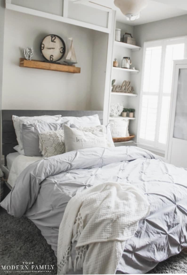 May include: A gray and white bedroom with a bed, a white blanket, and a wooden shelf with a clock and a sailboat. The shelf is above the bed. The bed has a gray headboard and a white comforter. The blanket is folded on the bed. The shelf is white and has a wooden top. The clock is round and has black numbers. The sailboat is white and has a blue sail.